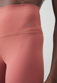 Aukštos liemens, prie kūno prigludusios leggingės iš minkštos koralinės medžiagos, su šviesiai pilku Nike logotipu juosmens srityje, pasižyminčios lygiu, matinio paviršiaus tekstūromis.