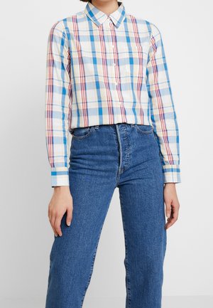 Kobieta ubrana w koszulę na długi rękaw w niebiesko-biało-czerwoną kratę, wpuszczoną w wysokie niebieskie jeansy, na jednolitym tle.