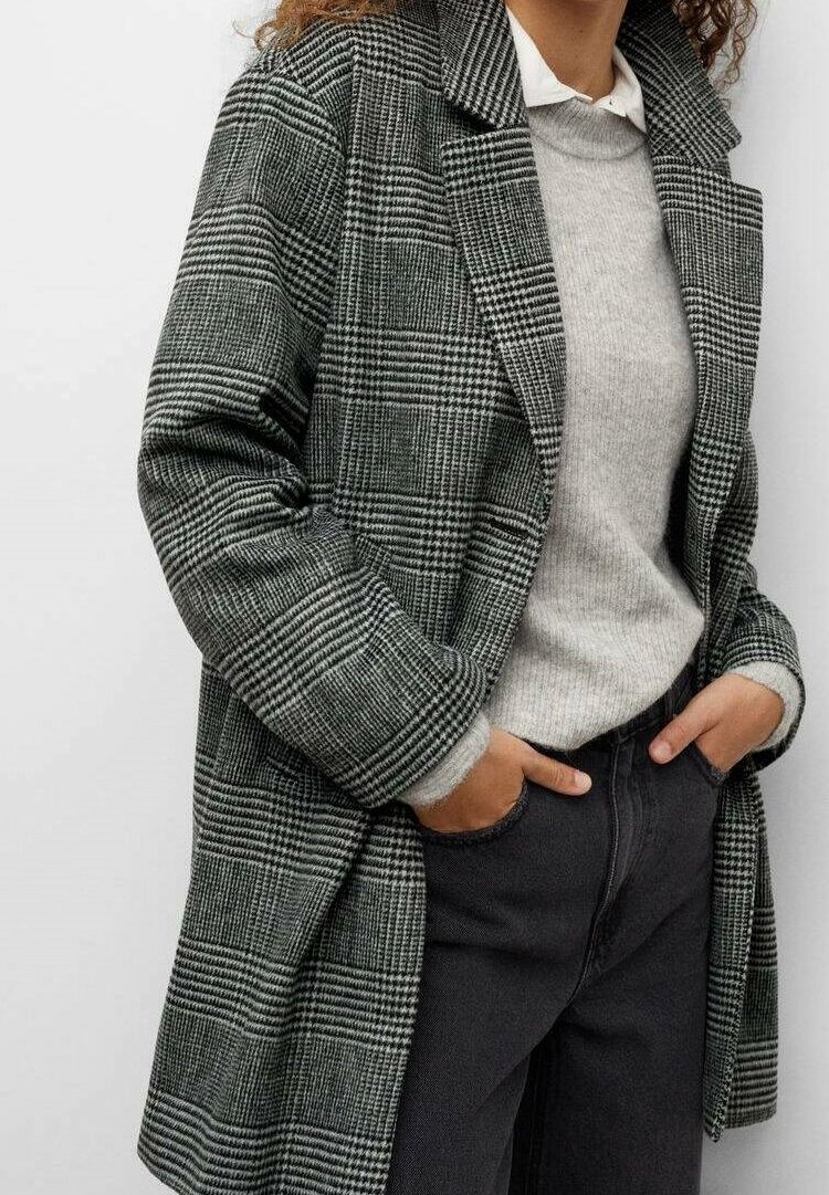 Personne portant un manteau à carreaux noir et blanc par-dessus un pull gris clair et un pantalon noir, les mains dans les poches, debout devant un fond blanc.