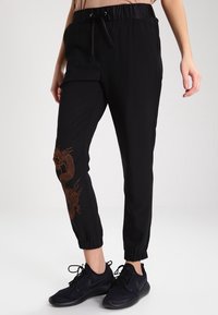 Black jogger pants with an elastic waistband and drawstring. Features an orange dragon design on the left leg and elastic cuffs at the ankles.