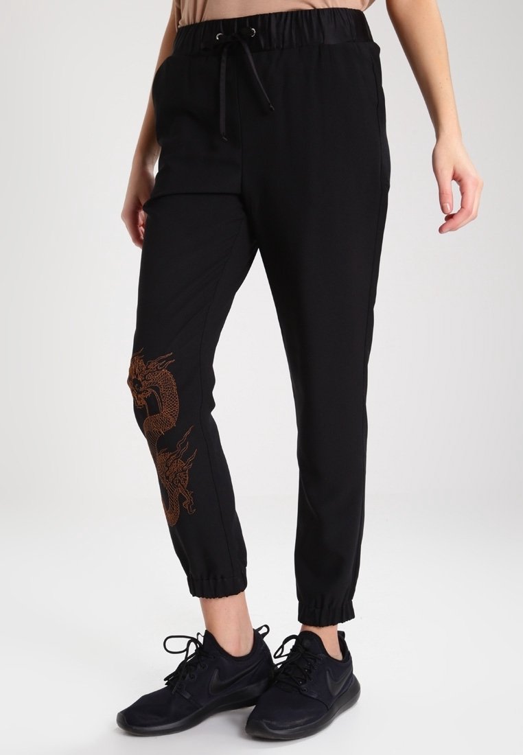 Black jogger pants with an elastic waistband and drawstring. Features an orange dragon design on the left leg and elastic cuffs at the ankles.