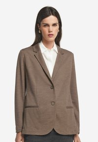 Brązowa marynarka z teksturowanej tkaniny, z wyłogowym kołnierzem, dwoma przednimi kieszeniami i dwoma guzikami. Noszona na białej koszuli.