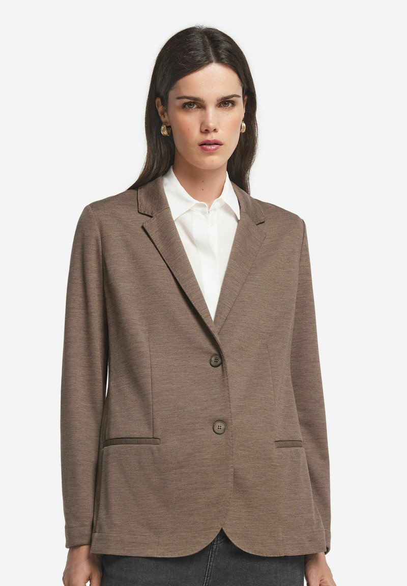 Brązowa marynarka z teksturowanej tkaniny, z wyłogowym kołnierzem, dwoma przednimi kieszeniami i dwoma guzikami. Noszona na białej koszuli.