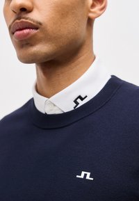 Κοντινό πλάνο ενός ατόμου που φοράει μπλε σκούρο πουλόβερ πάνω από ένα λευκό πουκάμισο με γιακά, με μαύρο και λευκό γεωμετρικό λογότυπο στον γιακά και στο στήθος.