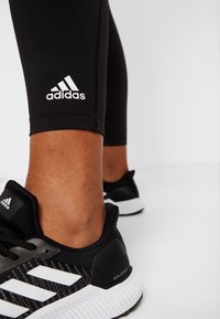 Mallas deportivas negras con un logo blanco de Adidas, combinadas con zapatillas de correr en negro y blanco de Adidas que presentan una textura de malla y rayas distintivas.