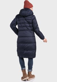 Marineblauer Puffermantel mit Kapuze, versehen mit einem gesteppten Design und einem langen Schnitt. Trägt eine dunkle Jeans und kastanienbraune Stiefel. Braune Strickmütze auf dem Kopf.
