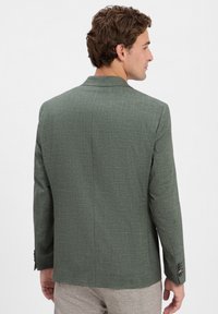 Männliches Model in einem stylischen grünen strukturierten Blazer, von hinten betrachtet, zeigt Details und Passform zusammen mit hellen Hosen.