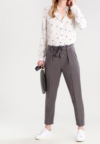 Biała koszula z guzikami i małymi czarnymi wzorami, szare spodnie z wysokim stanem i krawatem oraz białe trampki. Trzyma czarną torbę.