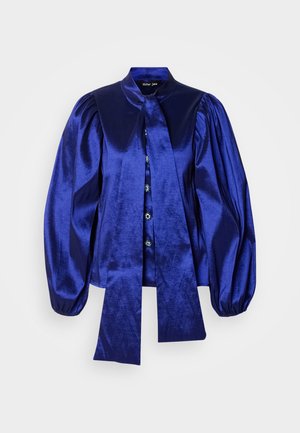 Szatén blúz sötétkék színben, puffos ujjakkal, gallérral és megkötős részlettel. Gombos záródás dekoratív gombokkal az elején.