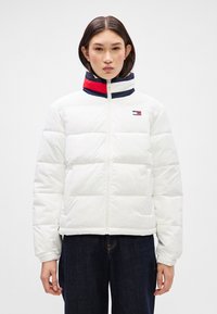 Balta pūkinė striukė su horizontaliai supjaustytais skydeliais, aukšta apykakle ir tamsiai mėlynos, raudonos ir baltos spalvų akcentu ties kaklu. Priekinis užtrauktukas.