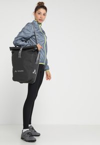 Zaino roll-top nero con accenti grigi, spallacci regolabili, tessuto impermeabile e fibbie laterali per la chiusura, indossato da una persona in abbigliamento sportivo.