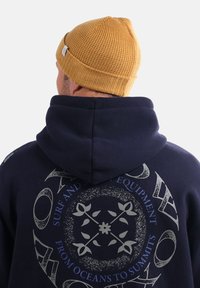 Sweat à capuche bleu avec un grand motif imprimé en gris et blanc au dos. Comprend un bonnet en tricot marron avec un motif texturé.