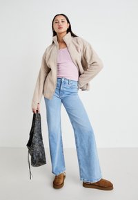 Beige fleecjakke, lys rosa tettsittende topp, lys blå vid bukse, brune slip-on sko, holder en svart veske med tekstur.