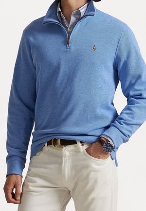 Hombre con un suéter de media cremallera azul claro sobre una camisa a rayas, pantalones blancos, cinturón marrón y un reloj de pulsera con esfera azul y correa de cuero marrón.