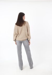 Mujer con cabello largo y oscuro, vestida con un suéter beige y jeans grises, de pie de espaldas contra un fondo blanco.