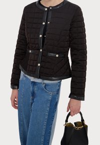 Veste matelassée noire avec un col rond, des finitions en cuir et une fermeture à boutons-pression. Accompagnée de jeans bleus et tenant un sac à main noir.