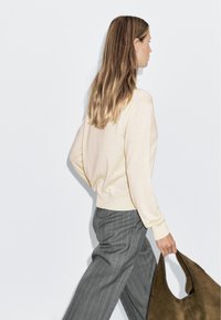 Pull léger beige à manches longues, associé à un pantalon gris à rayures. Le modèle tient un grand sac en daim brun.