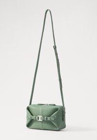 Borsa a tracolla in pelle verde con design a fiocco, hardware silver, e tracolla regolabile; presenta una chiusura con zip e dettagli cuciti.