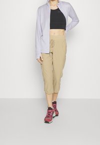 Giacca zip-up lilla chiaro, crop top nero, pantaloni beige a vita alta, e scarpe da trekking rosse con lacci rosa, che mostrano abbigliamento sportivo e funzionalità.