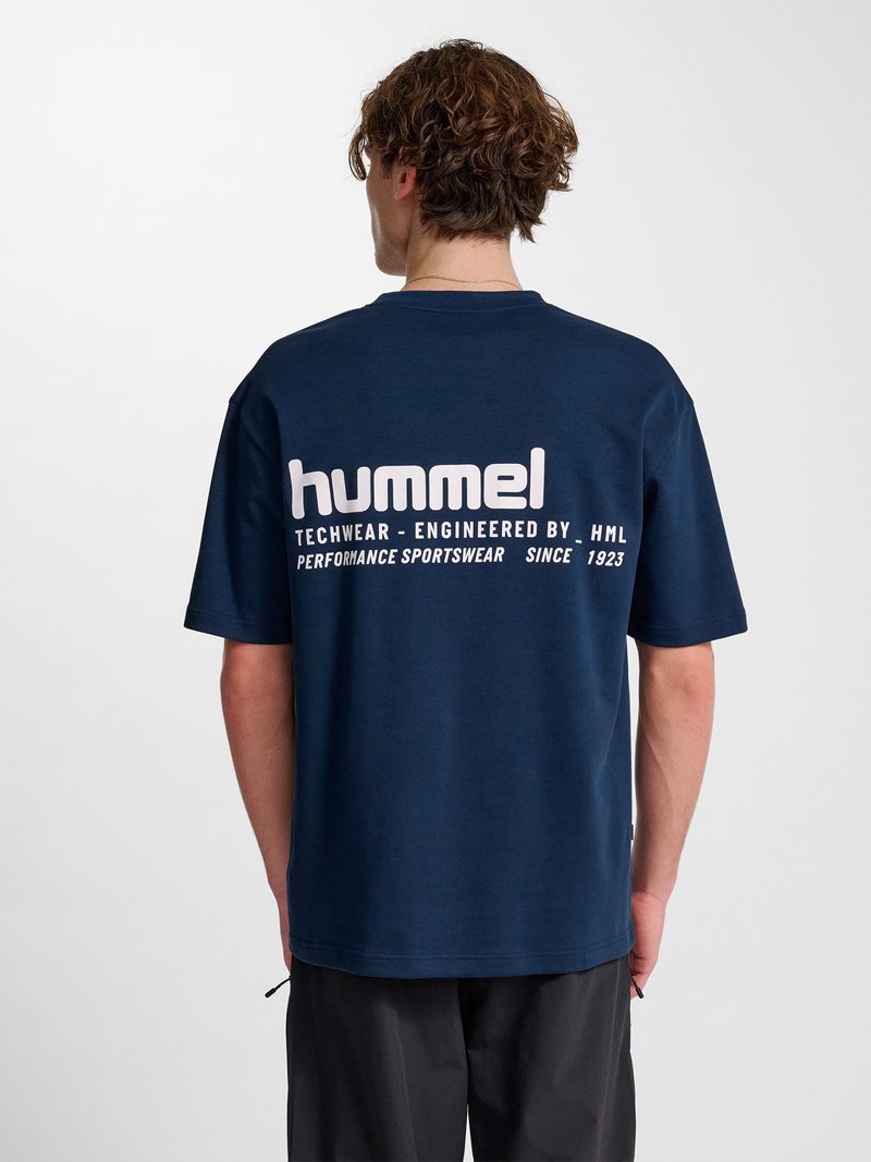 Mężczyzna o kręconych włosach, ubrany w granatowy t-shirt z białym napisem "hummel" i tekstem na plecach, stoi tyłem na jednolitym tle.
