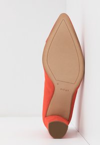 Chaussure à talon haut en daim rouge avec un embout pointu et une semelle extérieure lisse couleur tan. La semelle est équipée d'une texture antidérapante pour une meilleure adhérence.