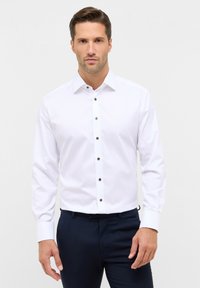 Weißes Langarmhemd mit klassischem Kragen, vorne mit braunen Knöpfen; kombiniert mit marineblauen Hosen. Glatte Stofftextur.