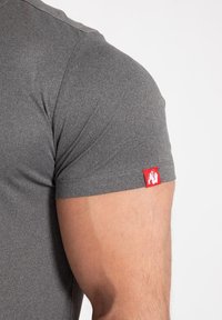 Siva kratka majica s kratkimi rokavi, izdelana iz gladkega materiala. Ima majhen rdeč logotip na rokavu, ki poudarja dizajn.