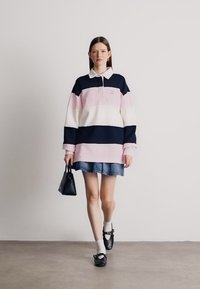 Ριγέ πλεκτό μπλουζάκι σε ναυτικό, ροζ και λευκό χρώμα, με γιακά και λογότυπο στο στήθος. Συνδυασμένο με τζιν φούστα και μαύρα παπούτσια, κρατώντας μια μικρή μαύρη τσάντα.