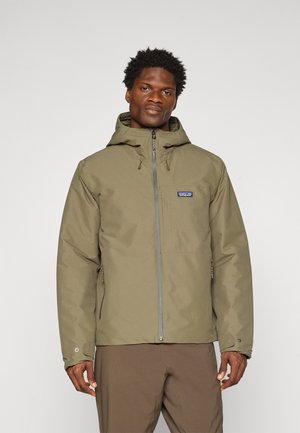 Man wearing olive green hooded jacket with front zipper and Patagonia logo, paired with brown pants, standing against plain background.