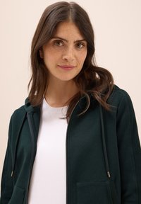 Felpa con zip di colore verde scuro realizzata in materiale morbido. Presenta un cappuccio con stringhe, tasche frontali e dettagli a costine sui lati.