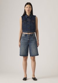 Gilet en denim au design court et avec des boutons, associé à un short en denim arrivant au genou. Les deux sont de couleur bleu moyen avec une texture décolorée. Des ballerines noires sont portées.