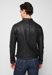 Svart läderjacka med en figursydd design, slät textur, subtila sömmar och tryckknappar. Bärs över mörkblå jeans, vilket visar bakifrån.