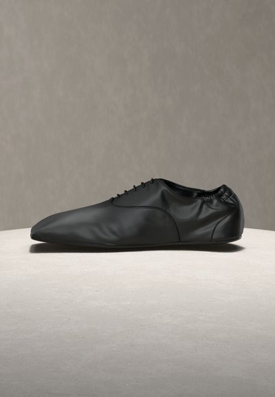 Chaussure pour homme en cuir noir à lacets, bout pointu et petit talon, présentée sur une surface grise avec un fond neutre.