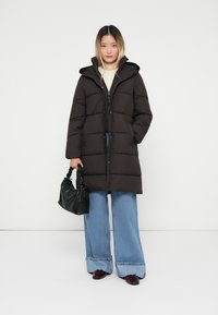 Černá péřová bunda s kapucí, má zip a patentní uzávěr. Modelka má na sobě široké modré džíny a drží malou černou kabelku.