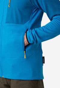 Giacca da performance blu con un motivo a griglia testurizzato, dotata di tasche con zip, un pannello a contrasto sulle spalle e polsini elastici.