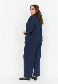 Femeie cu părul lung și brunet, îmbrăcată cu un top lejer albastru marin și pantaloni largi, cu pantofi negri, stând cu spatele la un fundal alb.