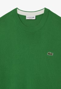 Pull vert à col rond avec col côtelé, arborant un petit logo crocodile Lacoste brodé sur la poitrine et une étiquette à l'intérieur de l'encolure.