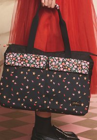 Černá taška s barevnými hvězdnými vzory, zdobená květinovými látkovými akcenty, pevnými držadly a zlatým kováním na zipu a logu.