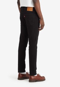 Jeans de denim negro con corte recto, que presentan una etiqueta de cuero en la cintura trasera y en los bolsillos traseros. Combinados con zapatos marrones y calcetines blancos.
