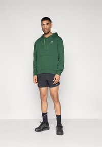 Πράσινο φούτερ με κουκούλα και μπροστινή τσέπη, με κορδόνια σε λάιμ πράσινο, σε συνδυασμό με μαύρο αθλητικό σορτς, μαύρες κάλτσες και μαύρα αθλητικά παπούτσια.