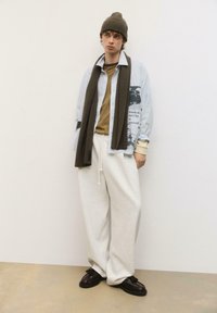 Model iført en stribet skjorte med knapper, graphic print, brun tørklæde, olivenfarvet beanie, beige sweatshirt og hvide sweatpants med mørke sko.