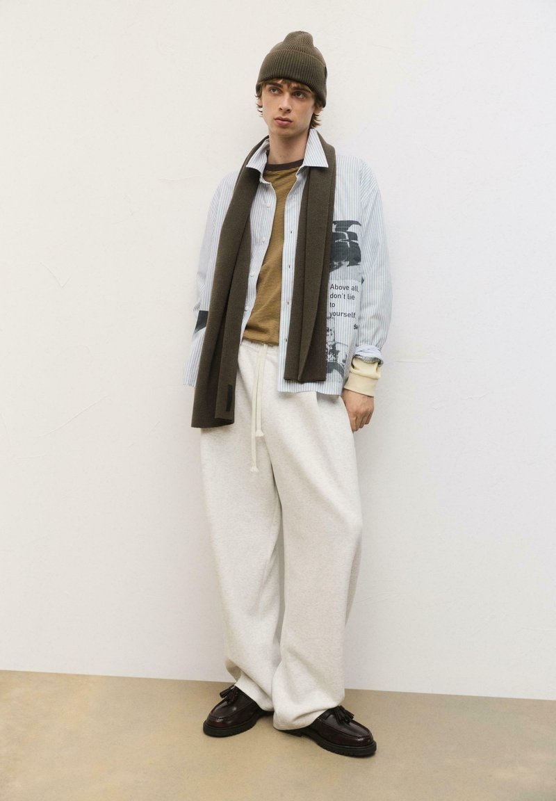 Model iført en stribet skjorte med knapper, graphic print, brun tørklæde, olivenfarvet beanie, beige sweatshirt og hvide sweatpants med mørke sko.