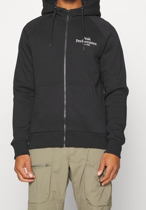 Person trägt einen schwarzen Kapuzenpullover mit Reißverschluss und weißem "Peak Performance est. 1986" Logo sowie beige Cargo-Hosen mit sichtbaren Taschen.