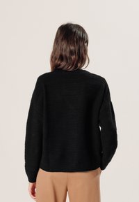 Schwarzer, strukturierten Pullover mit Rundhalsausschnitt und langen Ärmeln, der ein wellenförmiges Muster aufweist. Das Design ist locker geschnitten und am Saum gerippt.