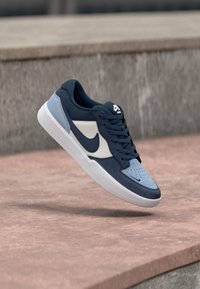 Sneaker navy și bleu deschis, cu un superior din suede și țesătură, logo-ul Nike alb, șireturi plate și perforații subtile pe zona de vârf.