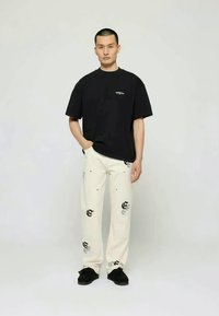 T-shirt noir à manches courtes avec logo, associé à un pantalon blanc avec des motifs imprimés noirs et des chaussures noires. Tenue simple et décontractée.