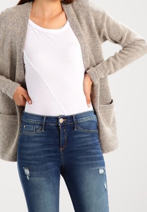 Femme portant un cardigan beige sur un haut blanc et un jean skinny bleu déchiré, les mains posées sur les hanches.