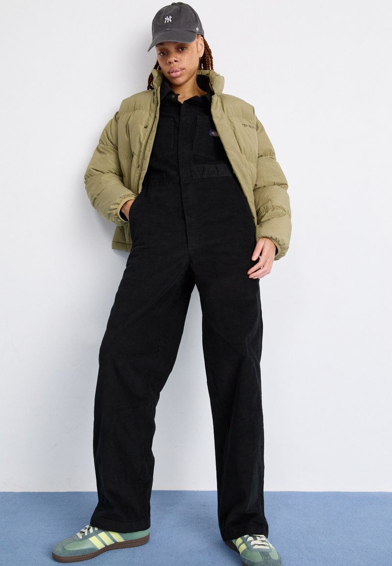 Veste pillow olive verte, salopette en velours côtelé noire, casquette de baseball grise et baskets vertes avec des rayures jaunes, présentant des tissus texturés.