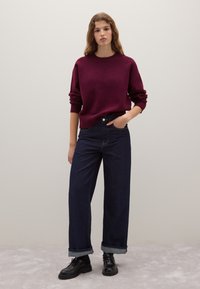 Bordeauxfarbener oversized Pullover mit umgeschlagenen Ärmelbündchen, kombiniert mit hochgeschnittenen dunkelblauen weiten Jeans und schwarzen Schnürschuhen.