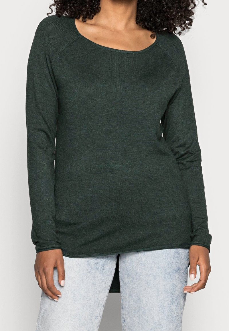 Femme portant un haut à manches longues vert foncé avec un col arrondi et un jean taille haute bleu clair, debout devant un fond uni.